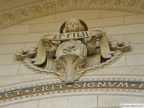 photo d'ange central du portail, aux traits féminins, portant un bannière nommant la seconde personne de Dieu dans le signe de croix : Et Filii